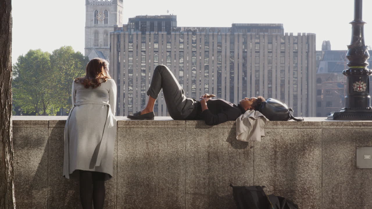 Black millennial woman lying on the embankment wall by the Thames in the City of London, talking with her friend, backlit