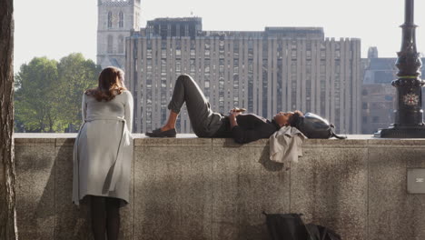 Black-millennial-woman-lying-on-the-embankment-wall-by-the-Thames-in-the-City-of-London,-talking-with-her-friend,-backlit