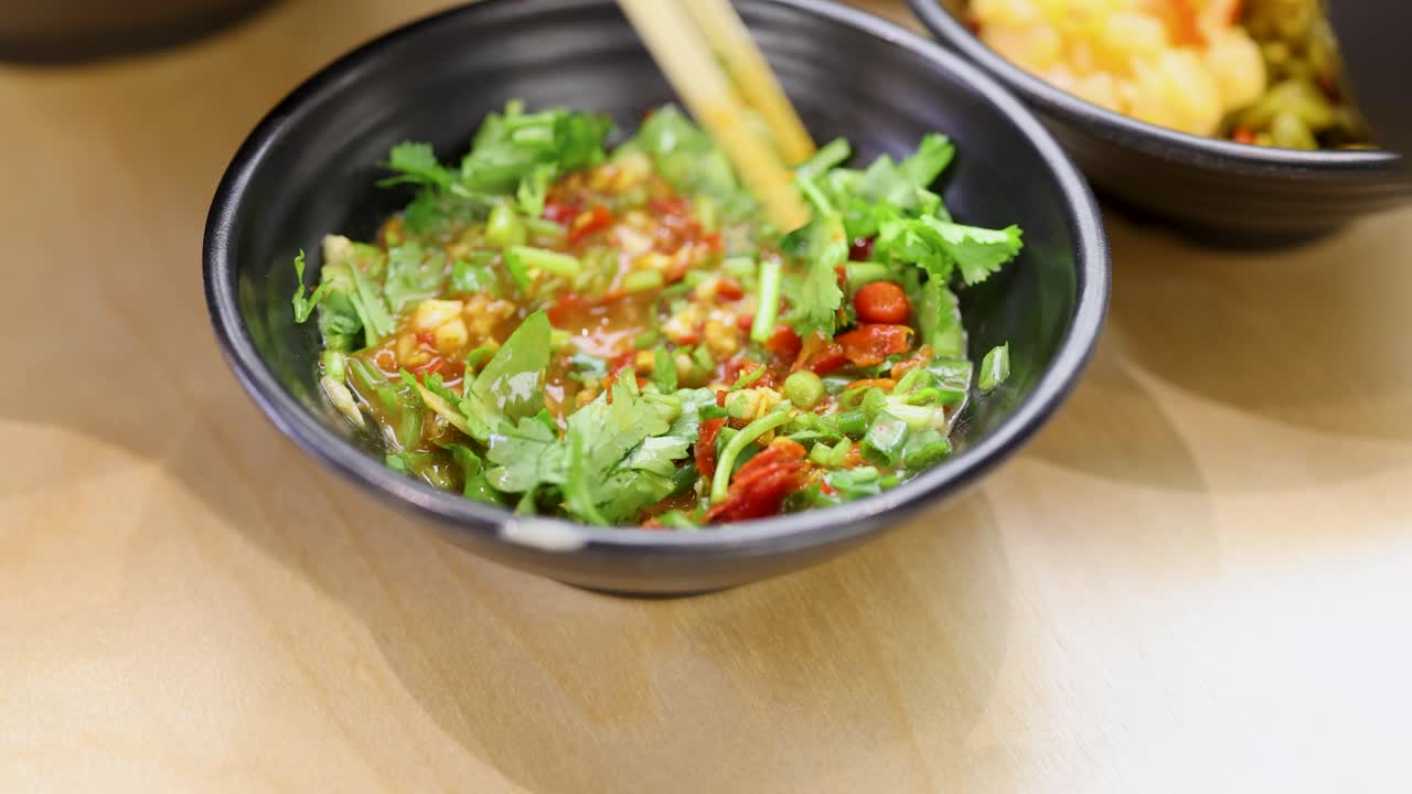 Chopsticks stir vibrant chili garlic sauce with cilantro, green onion, in black bowl, bright lighting