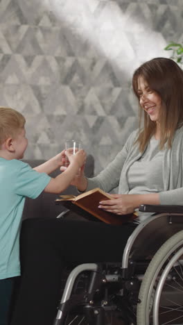 un niño cuidador trae un vaso de agua mientras su mamá con una lesión en la pierna lee un libro. un niño pequeño cuida a una mujer en silla de ruedas después de un accidente automovilístico