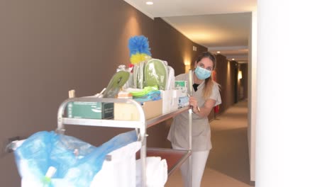 la criada con mascarilla y el carrito del piso caminan por un pasillo del hotel