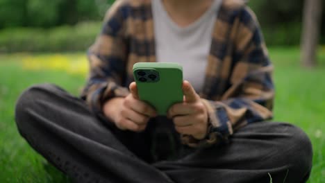 close up a girl in a plaid shirt and a white t-shirt is typing, shaking a green phone and chatting on social networks while relaxing in the park