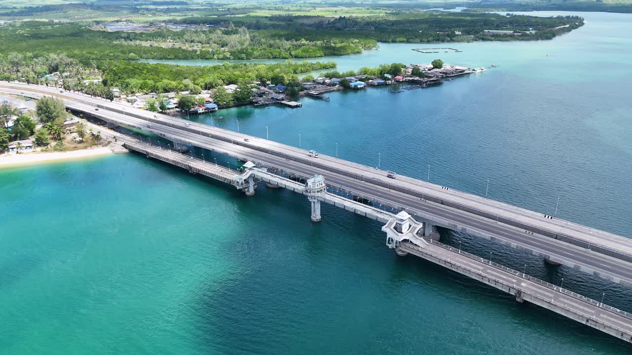 Drone footage captures Sarasin Bridge connecting lush landscapes over turquoise waters in Phuket, Thailand. Bright daylight enhances the vibrant scenery