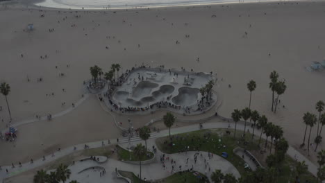 AERIAL:-Flying-towards-Venice-Beach-Skatepark-with-Visitors-and-Palm-Trees,-Sunset,-Los-Angeles,-California