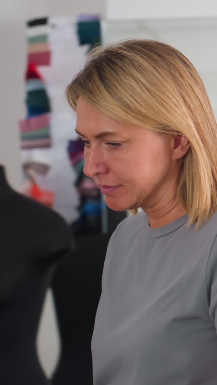 Mujer caucásica mirando el teléfono para comprobar las medidas, rubia con camiseta gris sentada en un estudio neutro, consultando el catálogo y enviando mensajes al proveedor, con expresión contemplativa mientras espera una respuesta