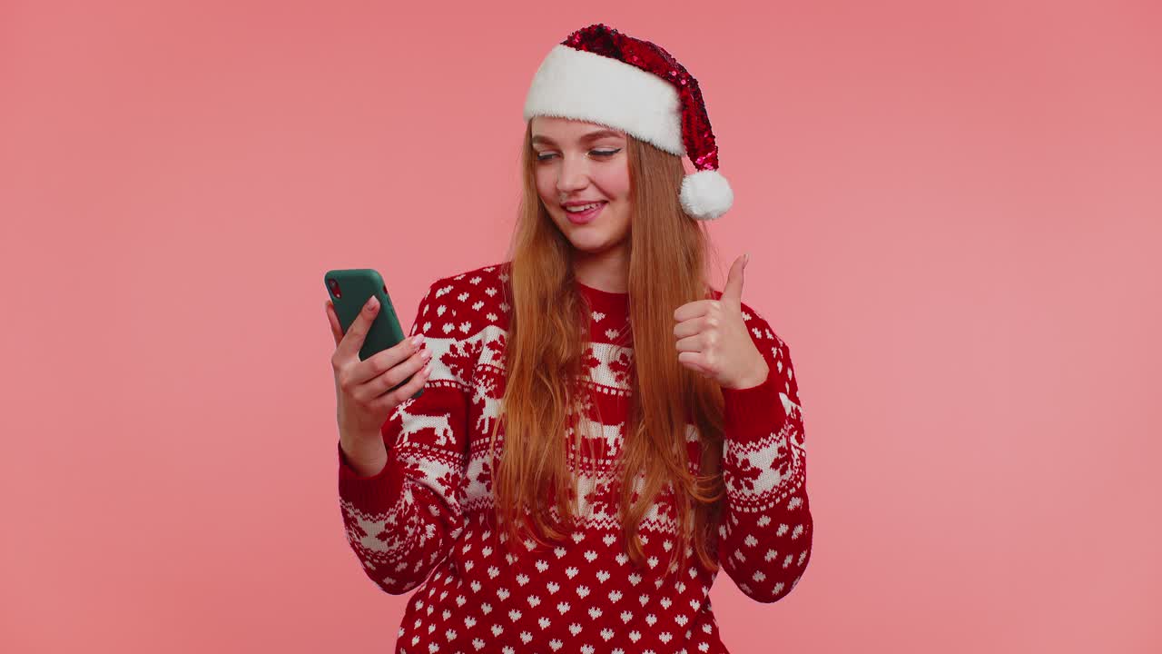 Woman in christmas sweater looking smartphone display sincerely rejoicing win success luck thumb up