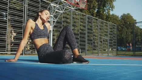 Fröhliches-Fittes-Mädchen-Mit-Airpods,-Das-Im-Sommersonnenlicht-Am-Sportplatz-Sitzt-Und-Sich-Nach-Dem-Training-Ausruht