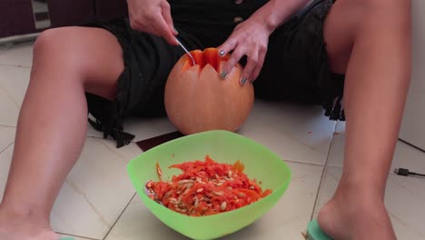 Girl-Sitting-Scraping-And-Removing-Seeds-Of-An-Orange-Pumpkin-For-Jack-o'-Lantern-Decoration