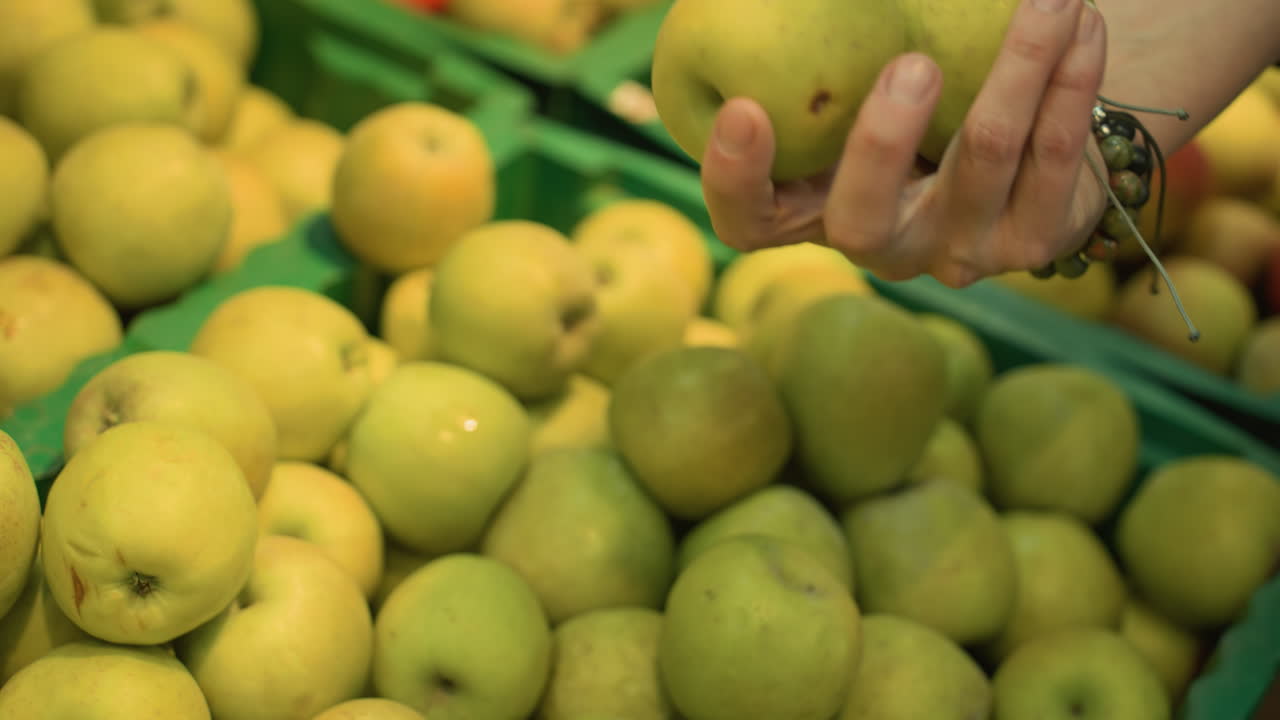 view of a hand wearing bead bracelets picking up two apples from a display in a grocery store. Shot with a handheld camera, capturing the detailed action in the produce section