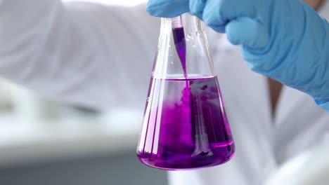 a hand in a blue glove holding a lab flask with purple liquid and a dropper
