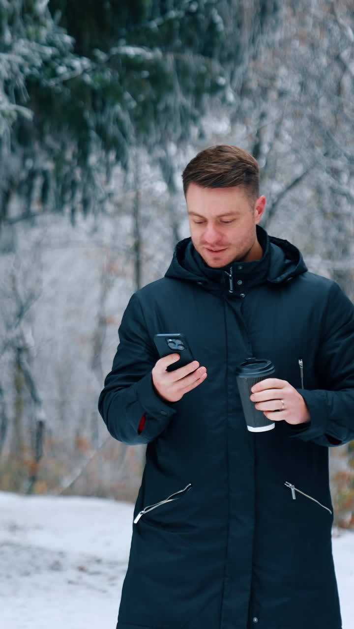 Confident male in black jacket looking at his phone. Man drinks coffee focused on his smartphone. Winter day at backdrop. Vertical video