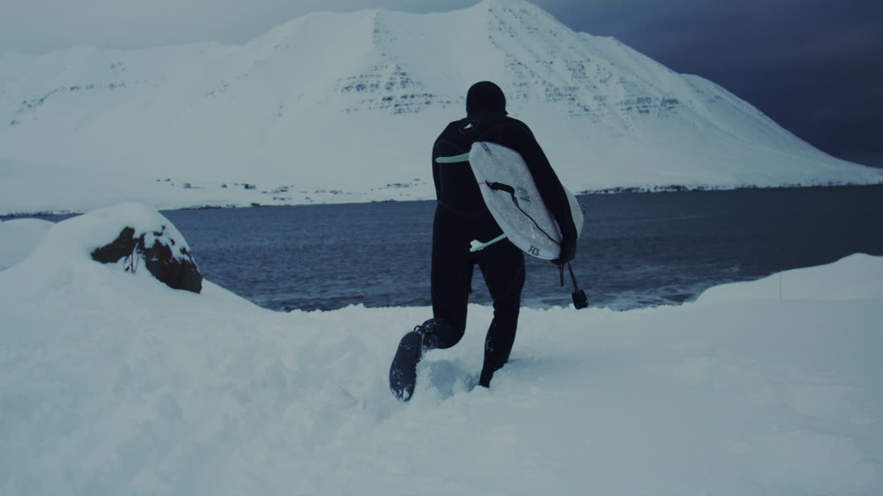Surfer stepping onto Arctic shoreline covered in snow under overcast winter skies, slow motion rearview