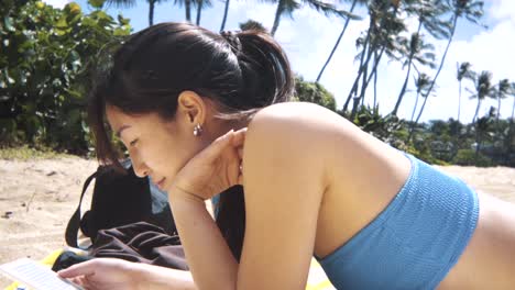 vista lateral de una chica en bikini azul acostada en la playa leyendo un libro de un lector electrónico