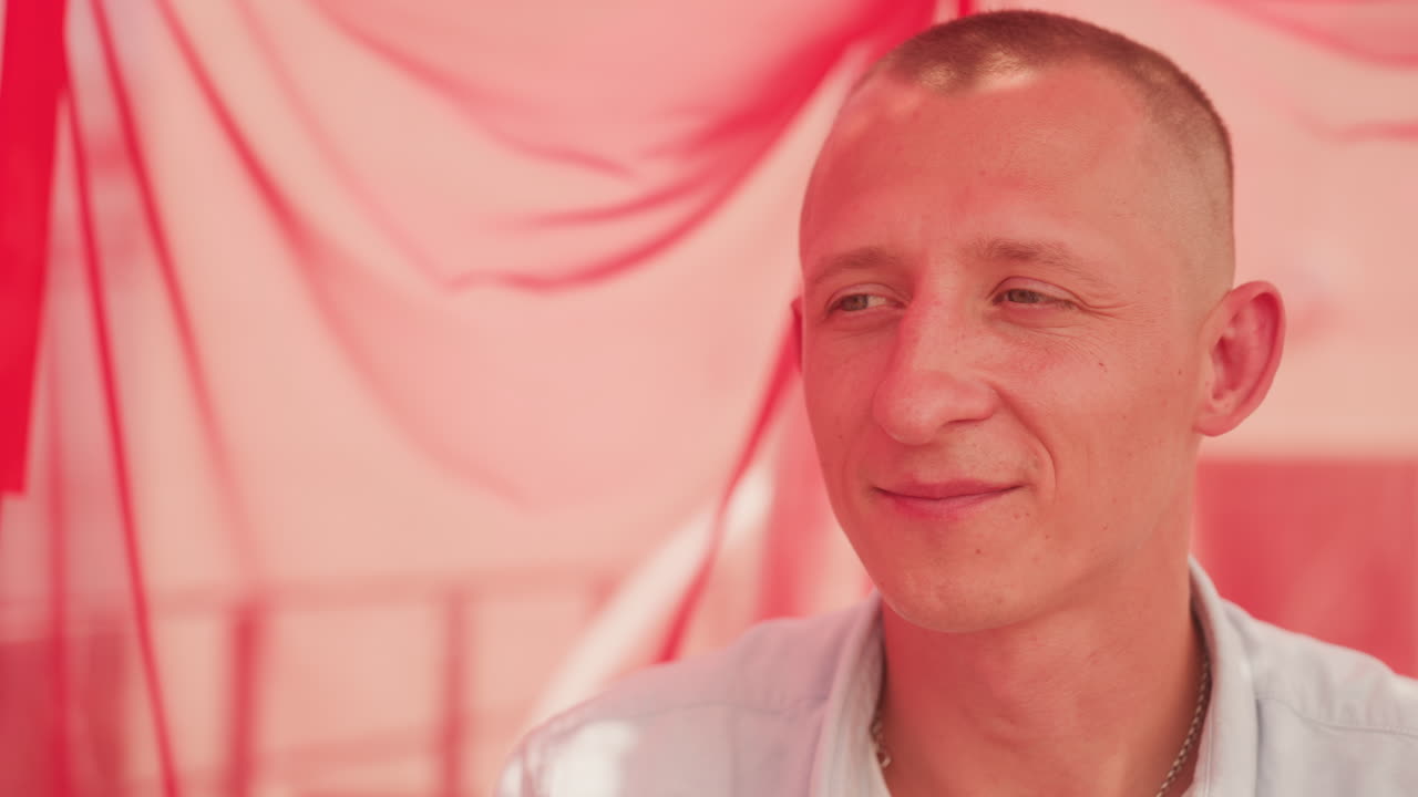 white man at red picnic canopy, intimate closeup of groom preparing romantic surprise, hand reaching for lighter, wicker basket and wine glass visible, warm pink lighting, contemplative smile