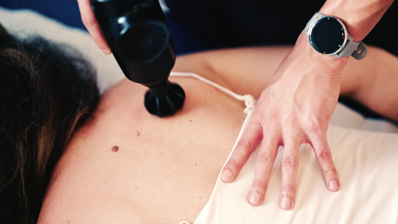 Therapist uses a percussion massage gun on a woman's shoulder while she lies on a treatment table