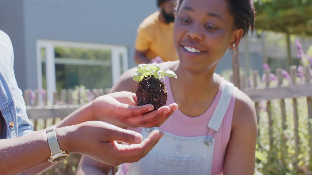 Adultos afroamericanos mostrando una plántula, niña con peto inspeccionando, aprendiendo jardinería en el jardín.