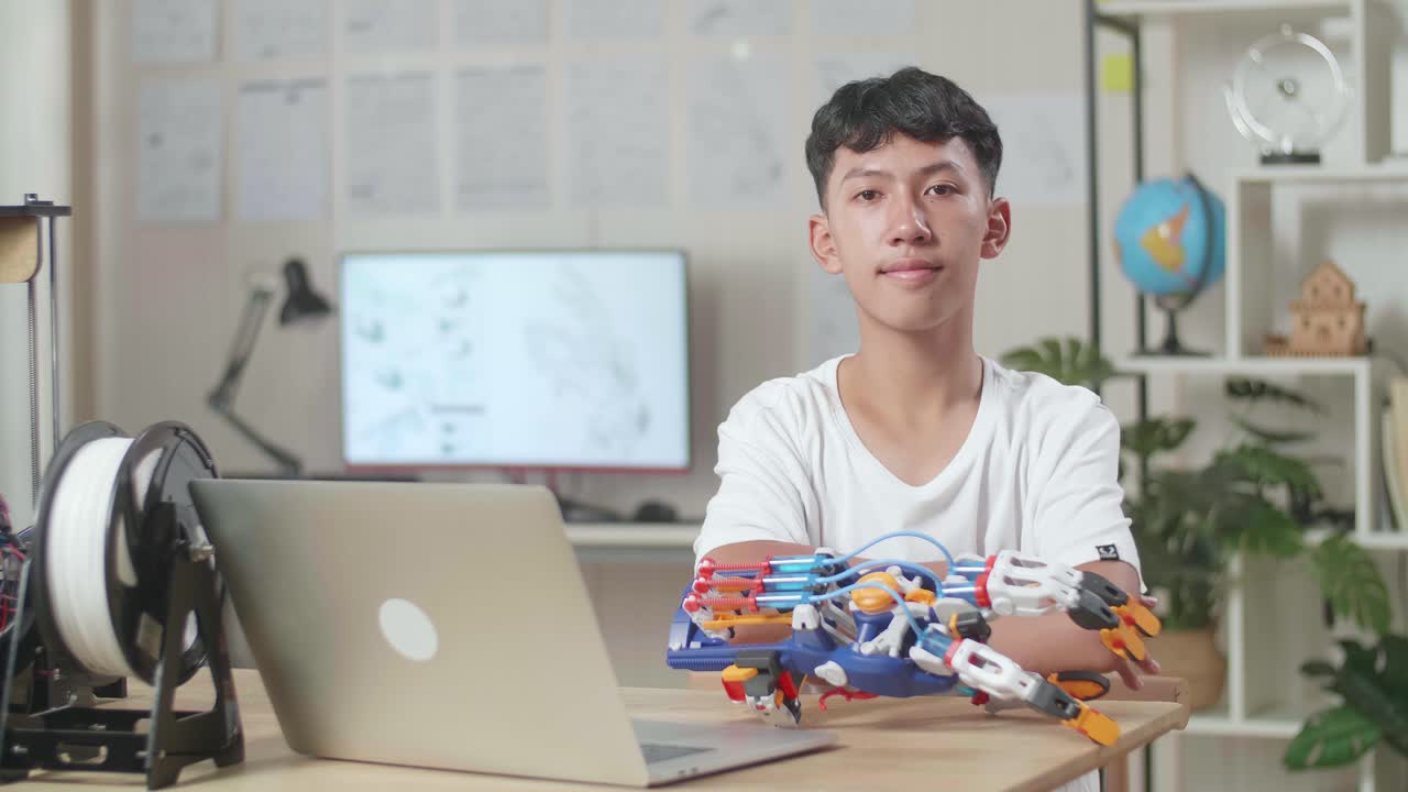 노트북을 들고 있는 아시아 소년은 집에서 사이보그 손에 대해 작업하는 동안 미소 짓고 카메라에 팔을 고 있습니다.
