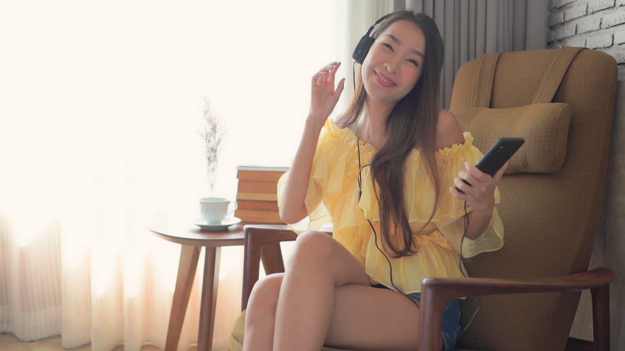 A happy young woman chair dances while listening to music through her headphones and smartphone