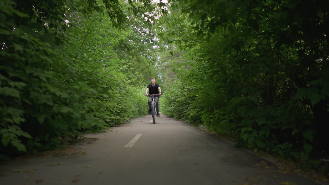 울창한 초록색으로 둘러싸인 길을 따라 자전거를 타고 다니는 어린 소년, 포장된 길에는 색 도로 표시와 건조한 잎이 어져 있으며, 소년은 어있는 가지를 피하기 위해 구부러집니다.