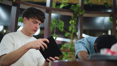 two friends at restaurant table, one snoozing face on crossed arms while companion taps phone screen, warm light bouncing off wood tabletop, background blur hinting menu and glassware soft ambience