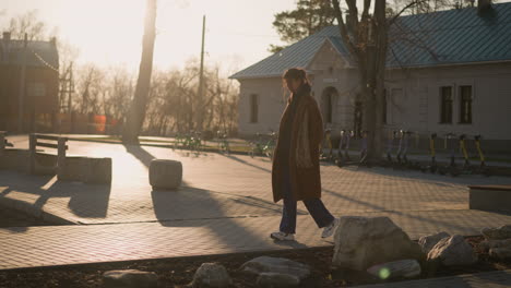 a girl wearing a brown coat, hoodie, and jeans wanders through a quiet park, looking disheartened. the scene is captured during sunset, with long shadows stretching across the pavement