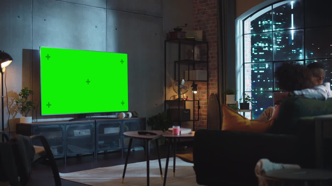 Black Couple In Love Sitting on a Couch, Watching Green Screen Chroma Key TV in Modern Apartment. Boyfriend and Girlfriend Have Tender Emotional Moments Together. Wide Side Back Shot