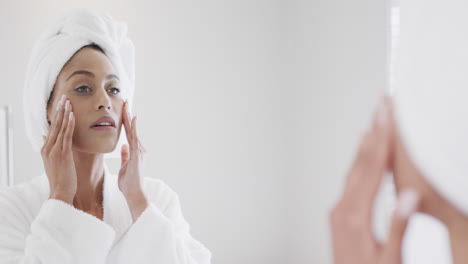 happy biracial woman massaging face looking in mirror in bathroom, slow motion