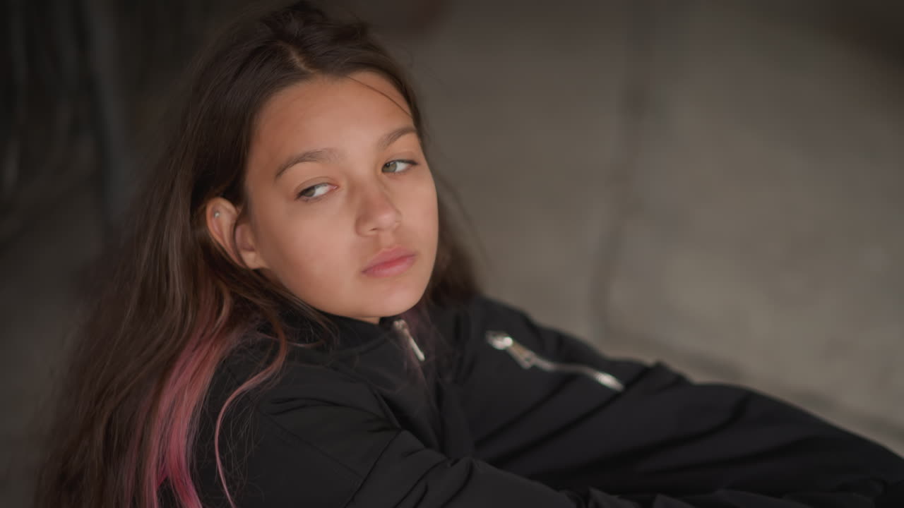 Adolescente latina seduta che riposa vicino a una ringhiera, scena pensierosa e intrisa di pioggia con la testa appoggiata sulle braccia, atmosfera intima e spontanea, ombra morbida, ciocca di capelli rosa, angolo urbano tranquillo, sottili dettagli di pozzanghere, sguardo introspettivo.