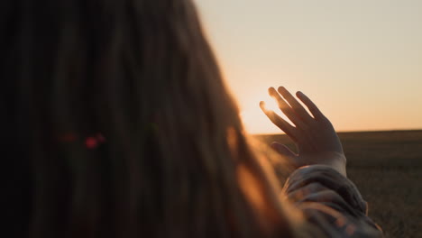 una chica con el pelo largo y hermoso extiende la mano hacia el sol poniente. vista trasera