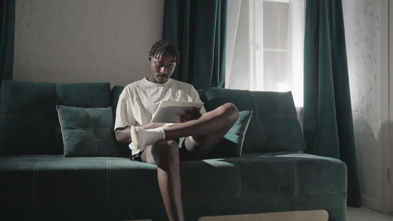 Champ sitting on teal sofa, relaxed, reading tablet with focus and calm demeanor. Soft sunlight filters through windows, creating a peaceful home environment with modern furnishings