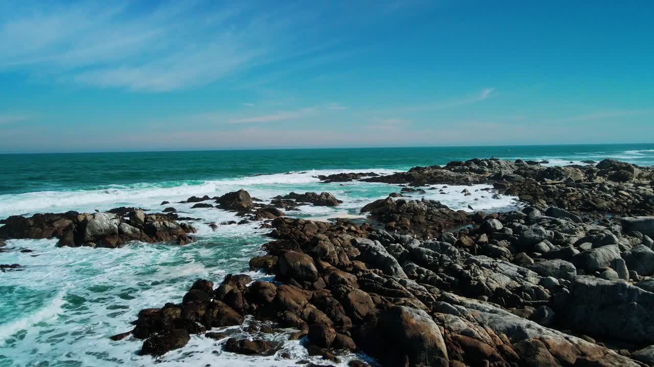 Ripresa con drone che sfiora una costa rocciosa cosparsa di massi con onde bianche schiumose che si infrangono tra rocce scure sotto un vivido cielo turchese del Pacifico. El Tabo, Cile.