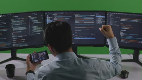 back view of happy asian man developer looking at code on mobile phone while write code with multiple computer screens in green screen studio