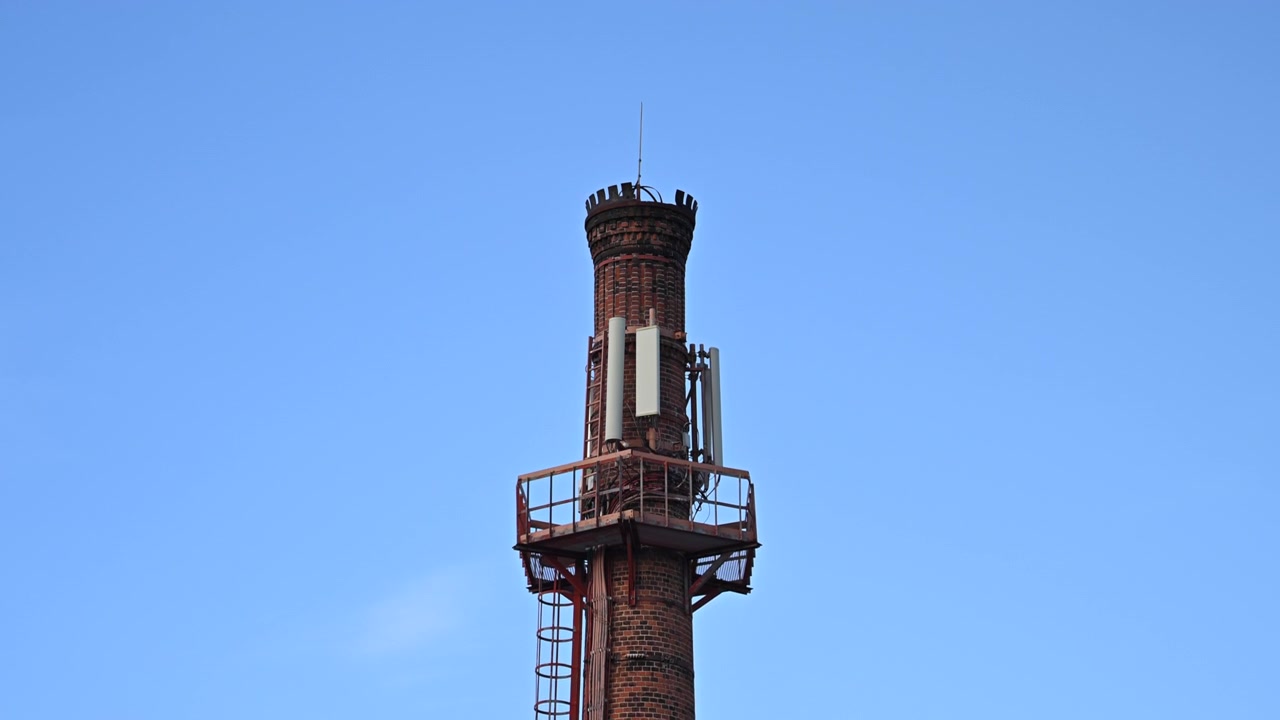 Primo piano di una storica ciminiera industriale in mattoni dotata di moderne antenne di comunicazione contro un cielo sereno.