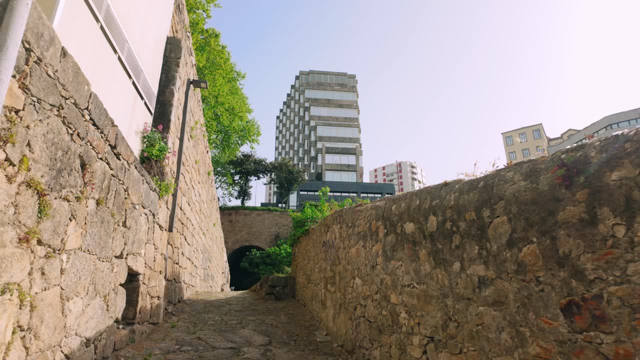 Stretta strada acciottolata che si snoda tra antiche mura in pietra in una giornata di sole a Porto. L'architettura moderna in lontananza contrasta con il fascino storico. Il fogliame verde incornicia il percorso.