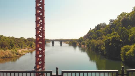 Drone-flying-through-the-red-Fair-Oaks-Bridge-sideways-with-a-view-of-the-American-River-and-Sunrise-Blvd-bridge---surrounded-by-green-trees---California