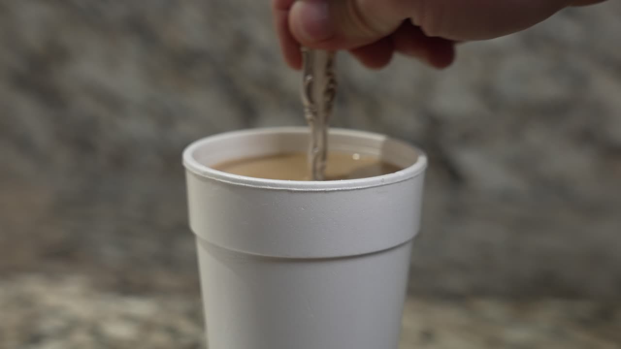 A man stirs their coffee with a silver spoon in a styrofoam cup