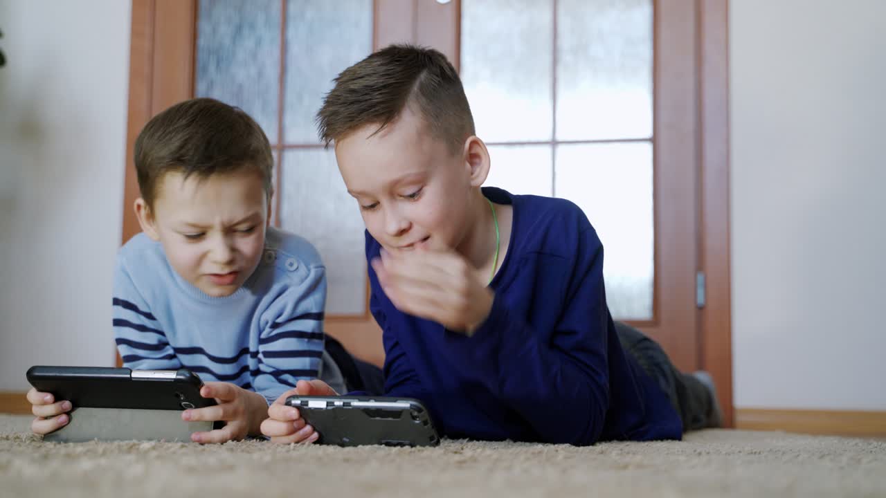 Two brothers involved in playing games on the digital gadgets at home. Excited boys lying on the floor and talking about enjoyable games on their electronic devices. Close-up
