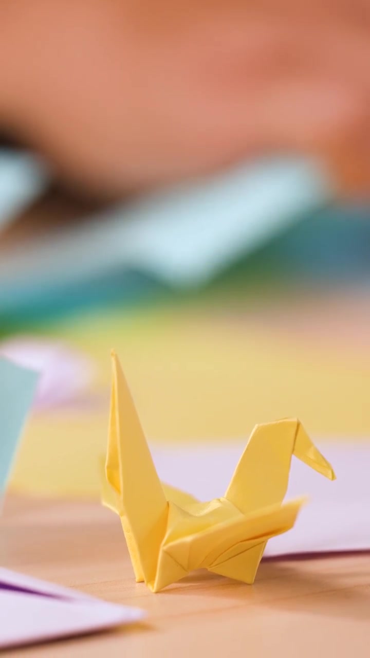 A yellow paper crane sits on a desk while hands fold origami in soft focus