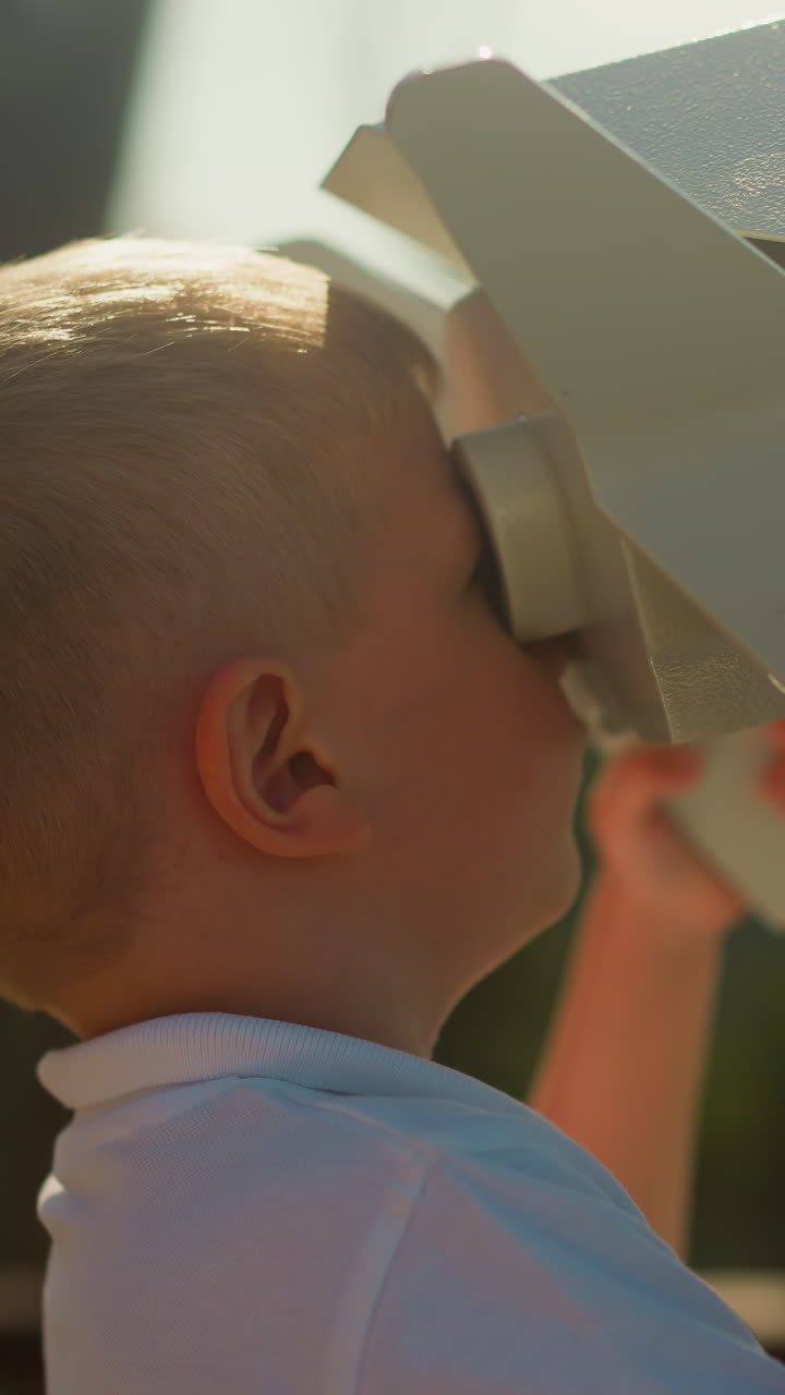 Inquisitive boy explores world via massive binoculars. Little child in white polo enjoys wonderful view of undulating mountains standing on observation deck
