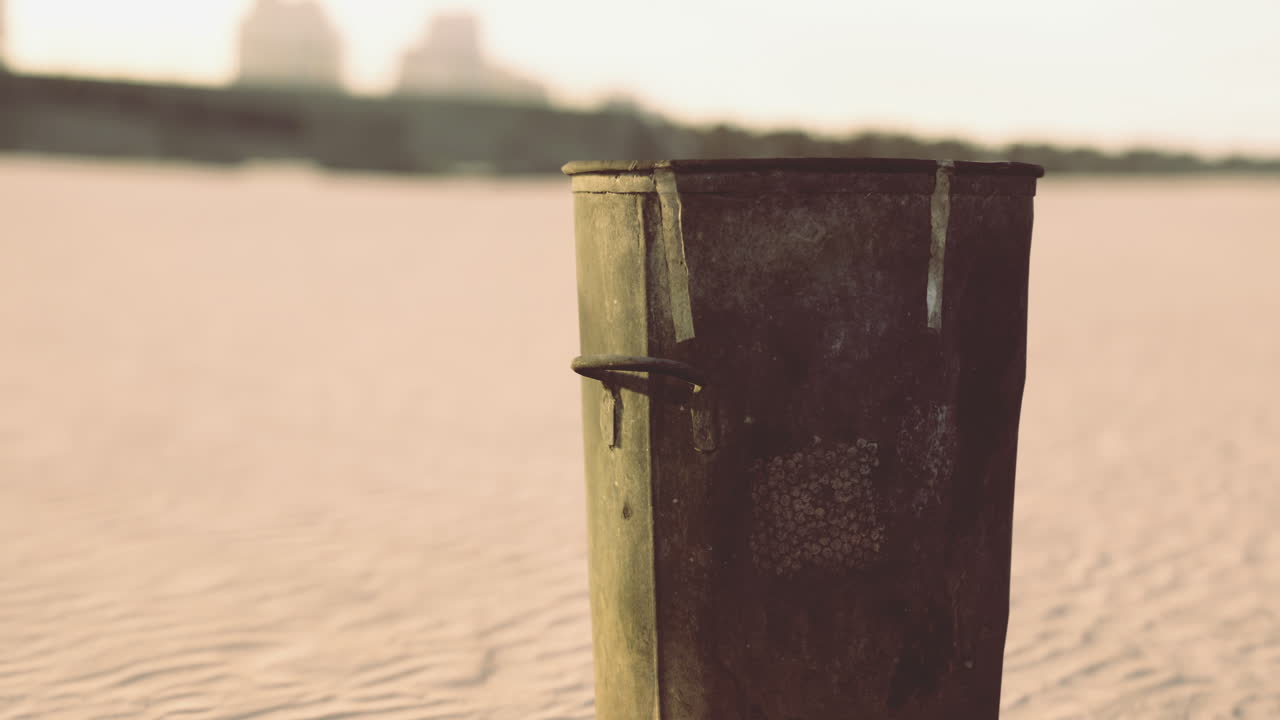A rusty trash bin stands on a sandy beach as waves gently lap the shore