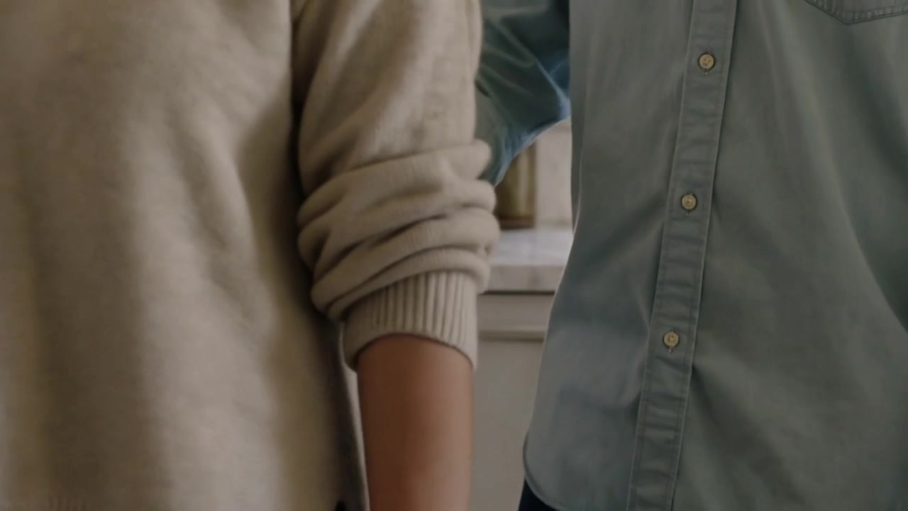 La telecamera si sposta, un uomo in camicia blu passa accanto e tocca una donna in maglione al bancone della cucina.