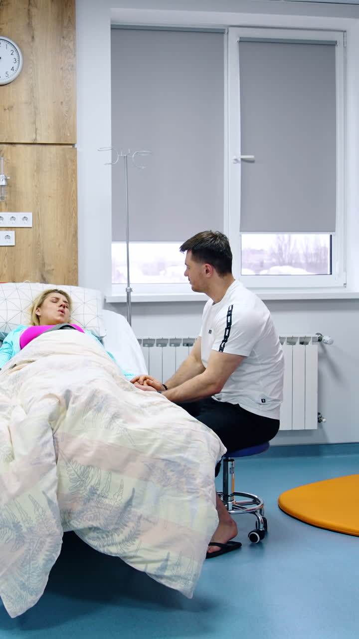 Loving husband sits at the bed of his pregnant wife. Blonde woman suffering from painful contractions. Modern hospital ward backdrop. Vertical video