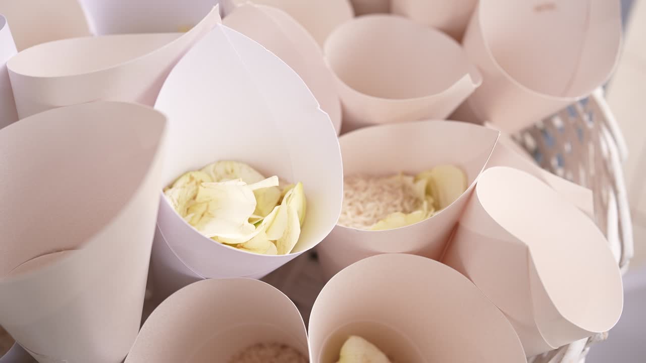 Pessoas estão fazendo cones de papel para guardar pétalas de flores em uma recepção de casamento. A atividade ocorre durante a tarde em uma área externa iluminada com mesas montadas.