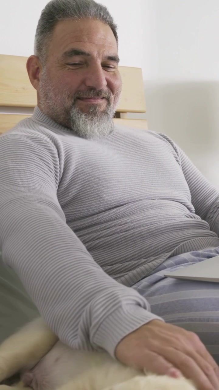 Video verticale: Uomo con maglione grigio che guarda il laptop e accarezza un cagnolino sul letto mentre naviga.