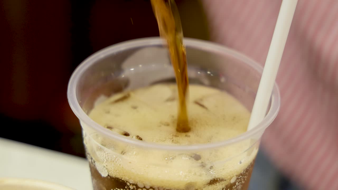 Hand pours dark carbonated soda from can into clear plastic cup filled with ice, creating foam. Bright indoor lighting, static close-up camera angle