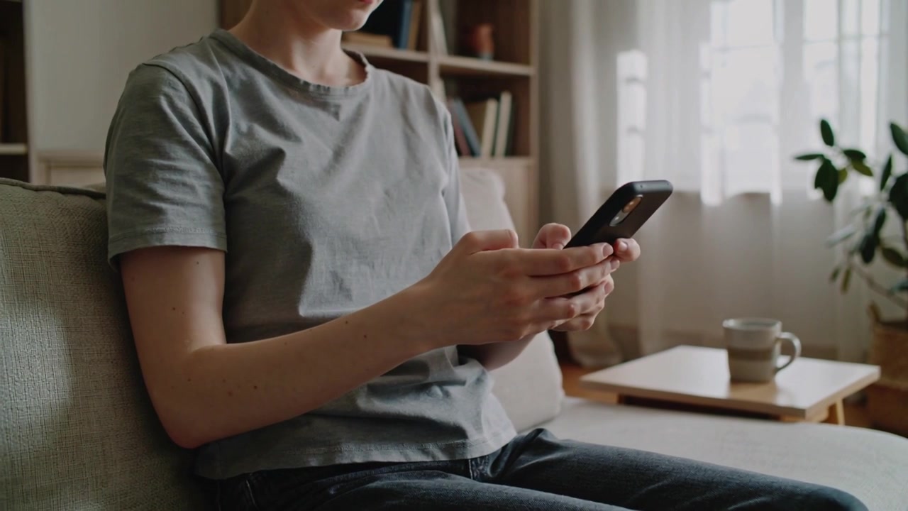 Žena v šedém tričku a tmavých džínách začíná s telefonem u klína a ťuká do smartphonu na pohovce v obývacím pokoji.