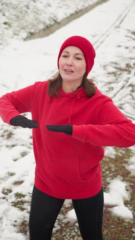 adult performing energetic winter workout outdoors lifting leg for balance on snowy ground near stairway wearing red hoodie and black gloves amid snowy hill pathway under foggy atmosphere