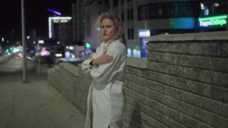 cold woman in white coat stands by city wall at night, arms crossed as wind gently sways her hair, she looks contemplative with hotel and residential buildings illuminated in background
