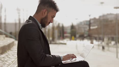 focused man working with laptop on knees while sitting in park