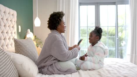 Happy-african-american-mother-combing-daughter-hair-sitting-on-bed-in-bedroom,-slow-motion